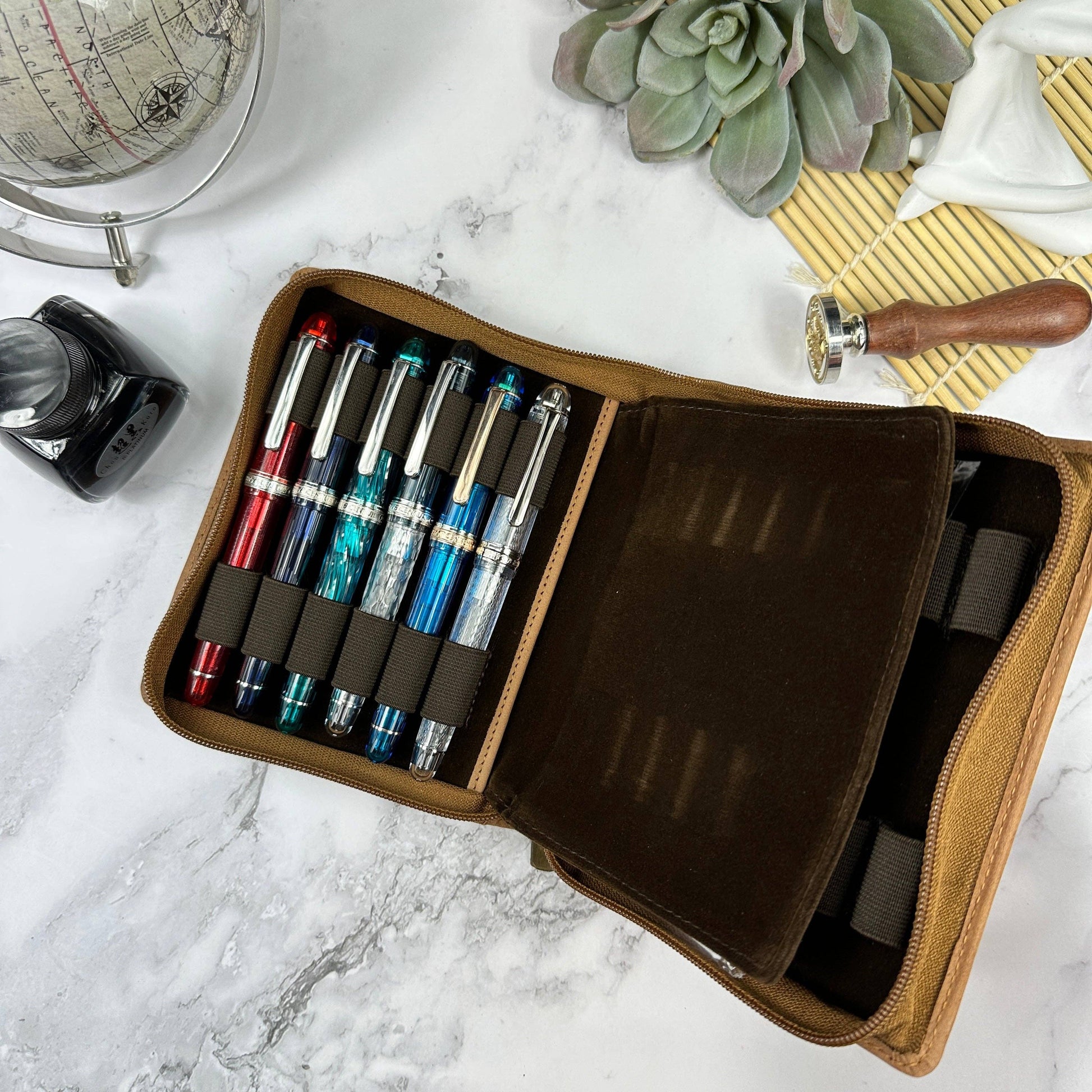 Set of fountain pens in a Girologio 12 pen leather case on a marble surface with a globe and plant in the background.