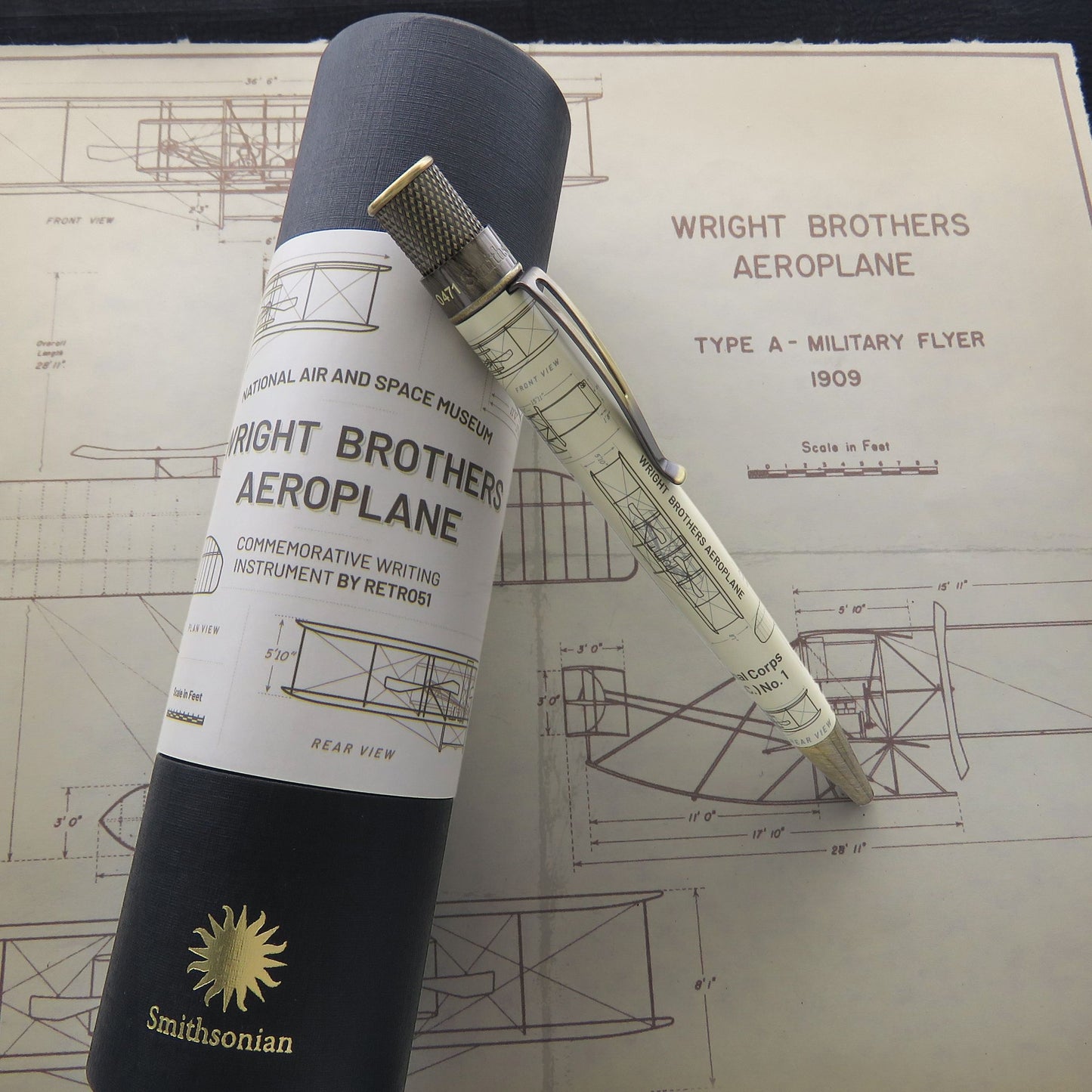 A commemorative writing instrument pen with Wright Brothers Aeroplane blueprint graphics, shown resting on top of its dark gray Smithsonian-branded cylindrical presentation tube, with a Wright Brothers Aeroplane print in the background.
