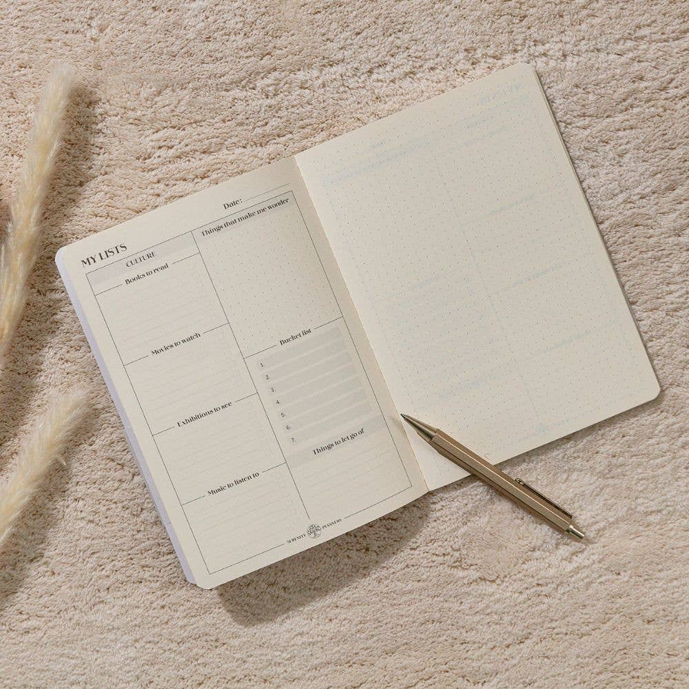 An open Clairefontaine_Bucket_List_Planner_A5_white grid planner or notebook with a gold pen resting on it, photographed from above on a textured beige fabric background with dried grass stems at the side.