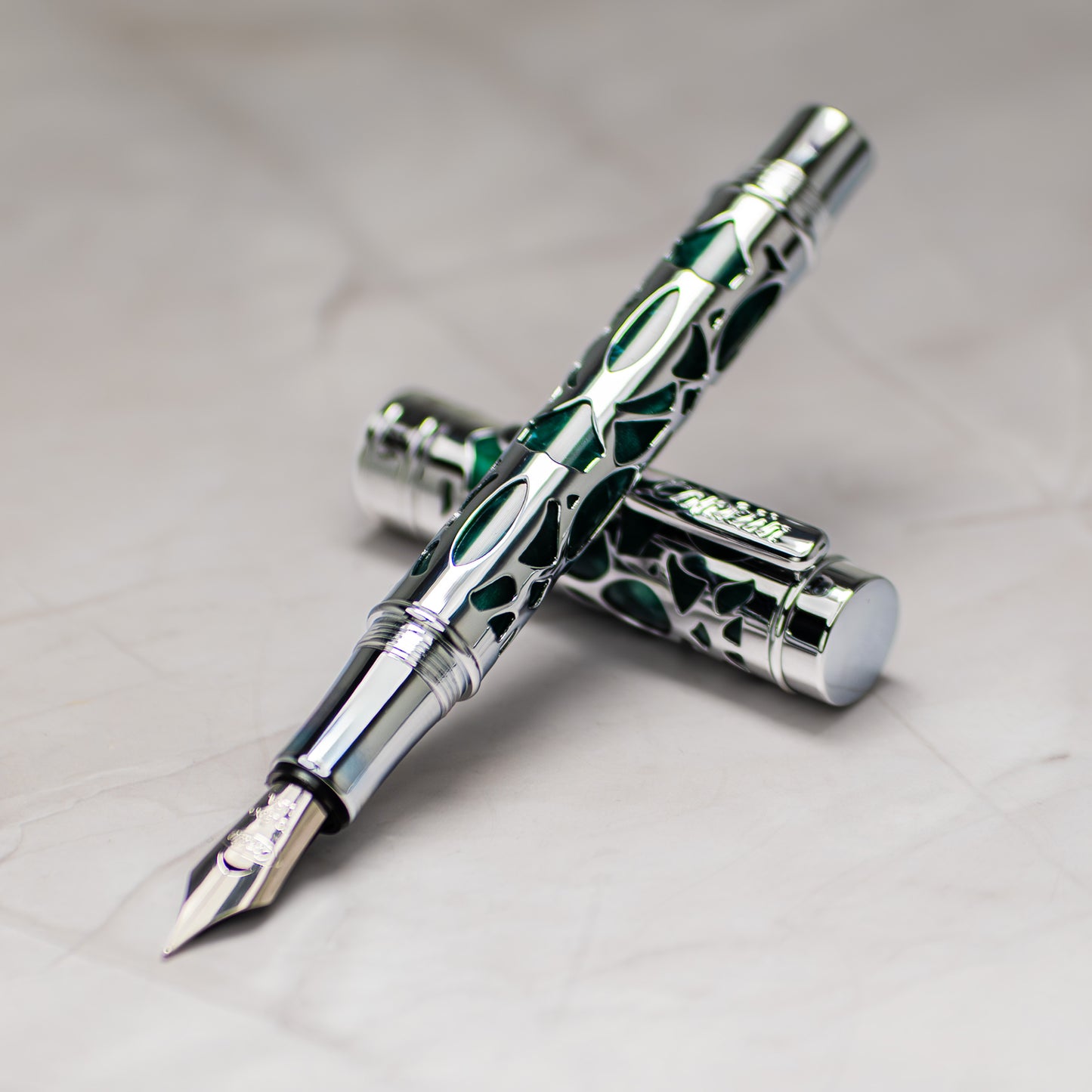 A close-up of a Conklin Endura Deco Crest Green fountain pen with a green and silver patterned barrel and a silver nib, shown with its cap placed behind it on a light marble-like surface. 