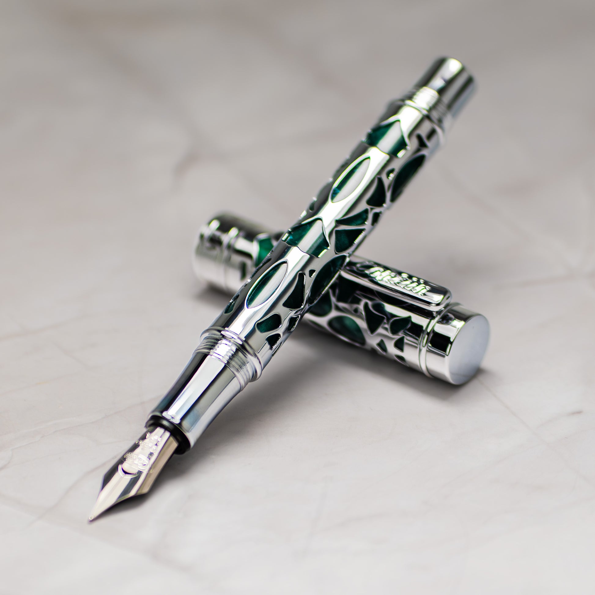 A close-up of a Conklin Endura Deco Crest Green fountain pen with a green and silver patterned barrel and a silver nib, shown with its cap placed behind it on a light marble-like surface. 