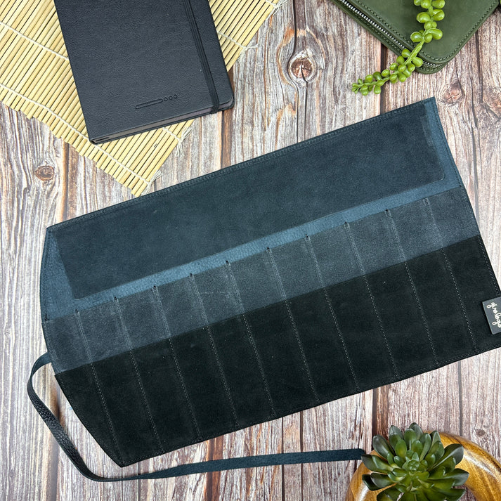 Black and gray striped pen roll bag on a wooden surface with a notebook and plant in the background