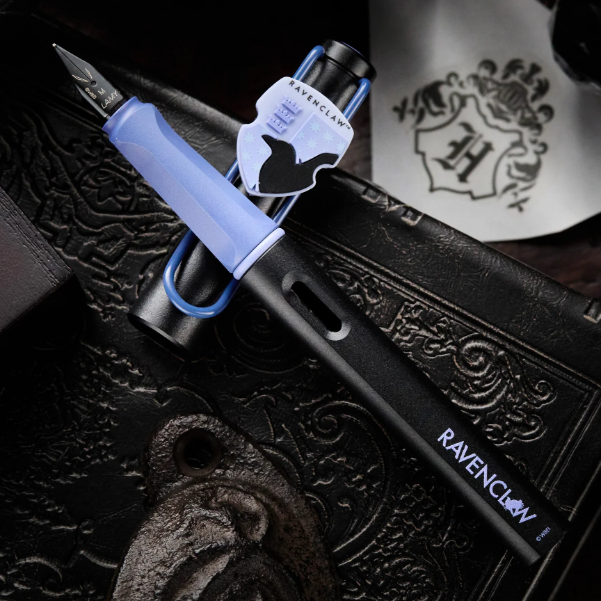 A Ravenclaw fountain pen with a black body, light blue grip, and silver nib, resting on a dark embossed leather surface with a Ravenclaw crest sticker nearby.