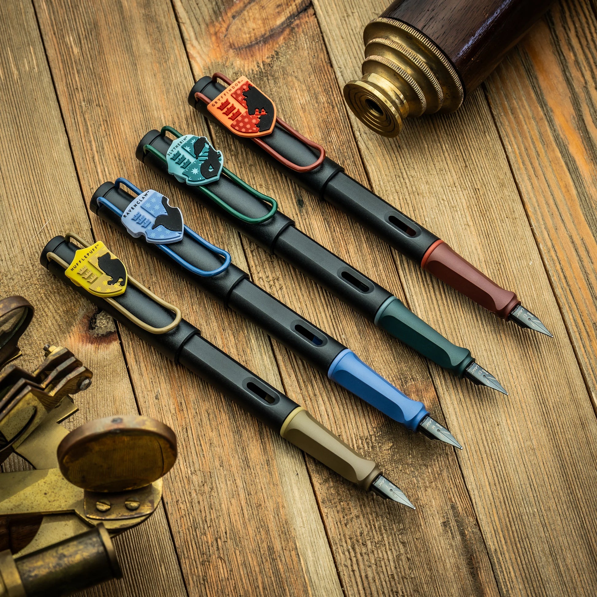 Four LAMY fountain pens with shield-shaped clips in different colors, arranged diagonally on a rustic wooden surface with vintage brass and wood-toned props.