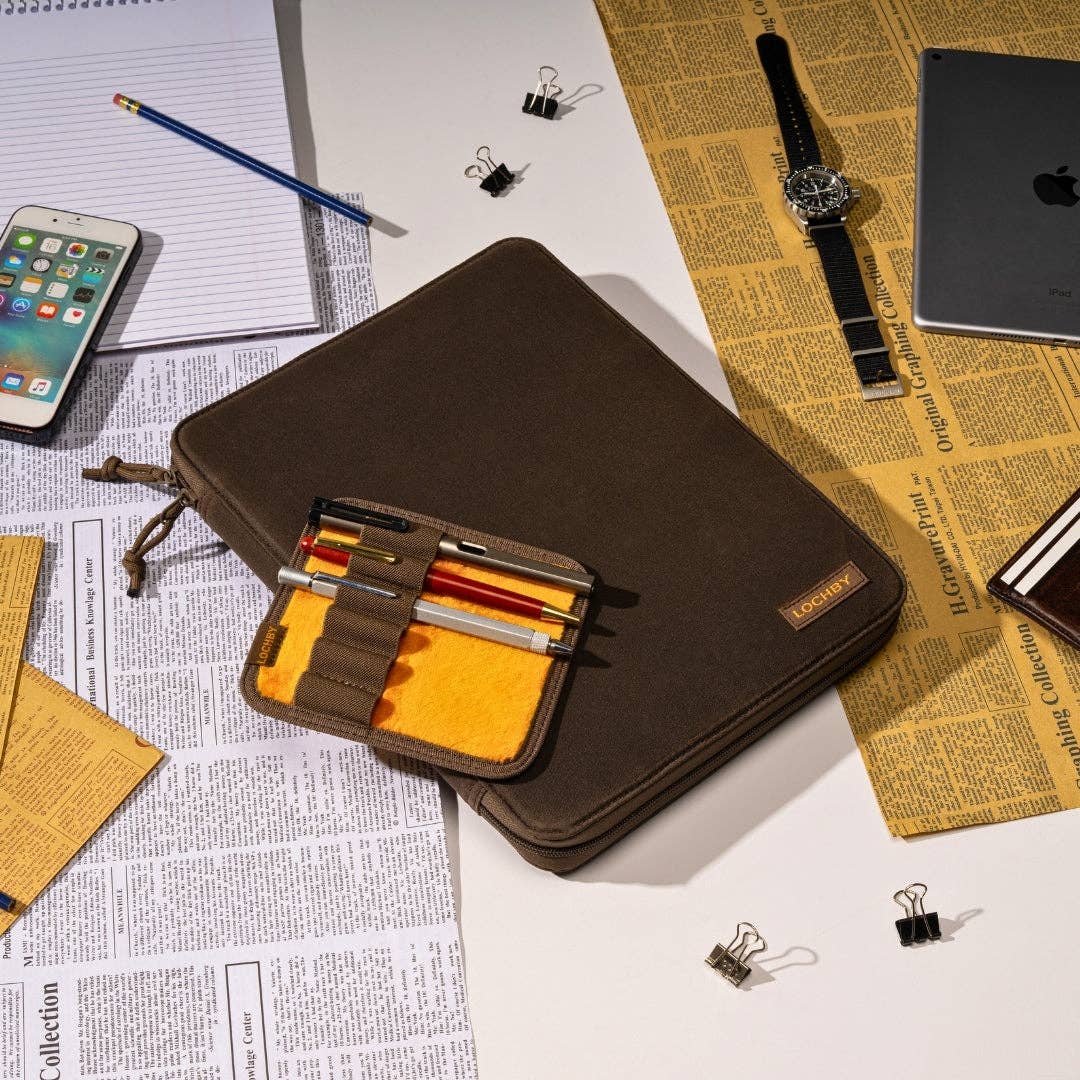LOCHBY Daily Folio A4 A brown laptop sleeve with a matching pen organizer pouch placed on a desk surrounded by office props including a phone, notebook, pencil, watch, laptop, and binder clips on newspaper print.