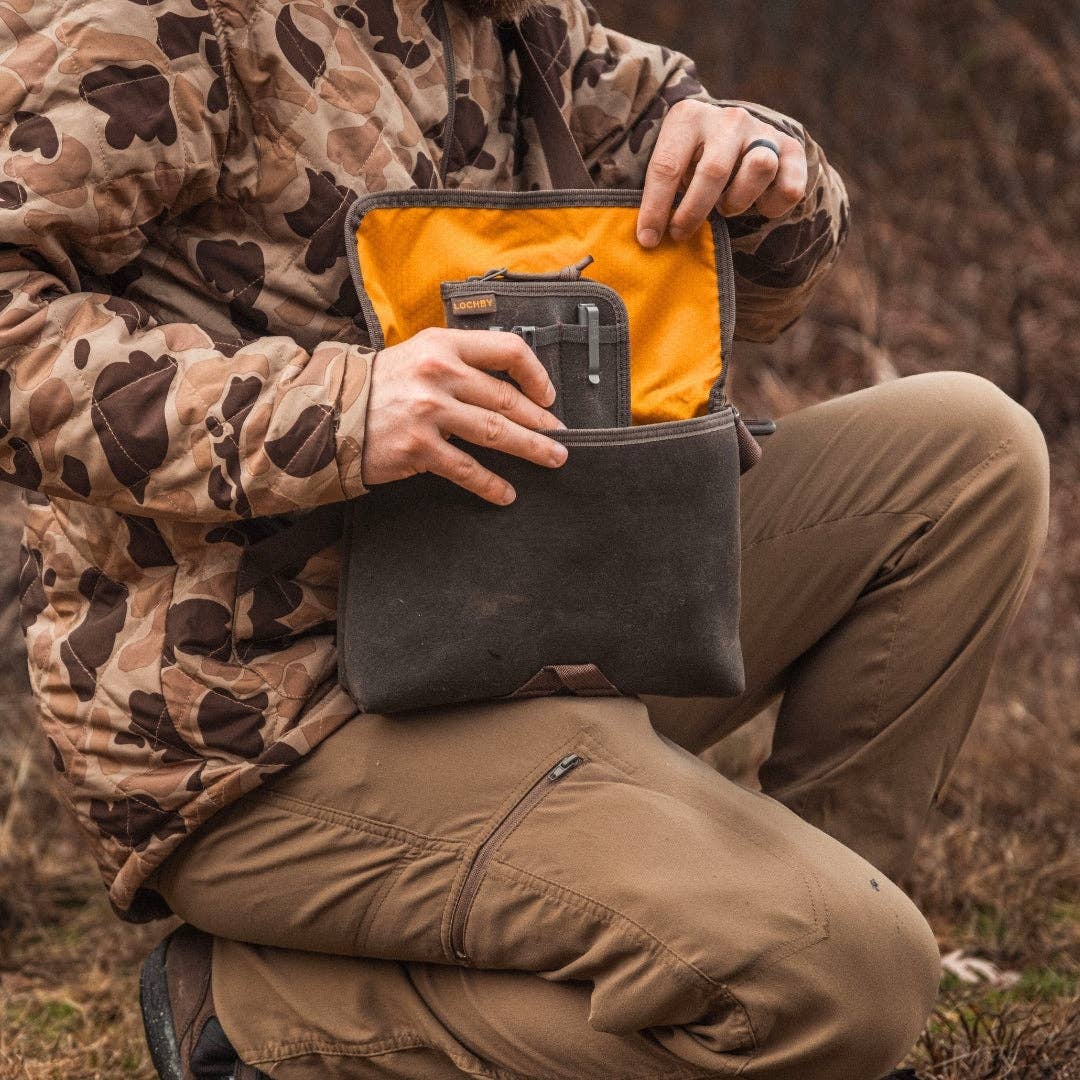 Person in camouflage jacket and brown pants holding a LOCHBY field sling with an orange interior.