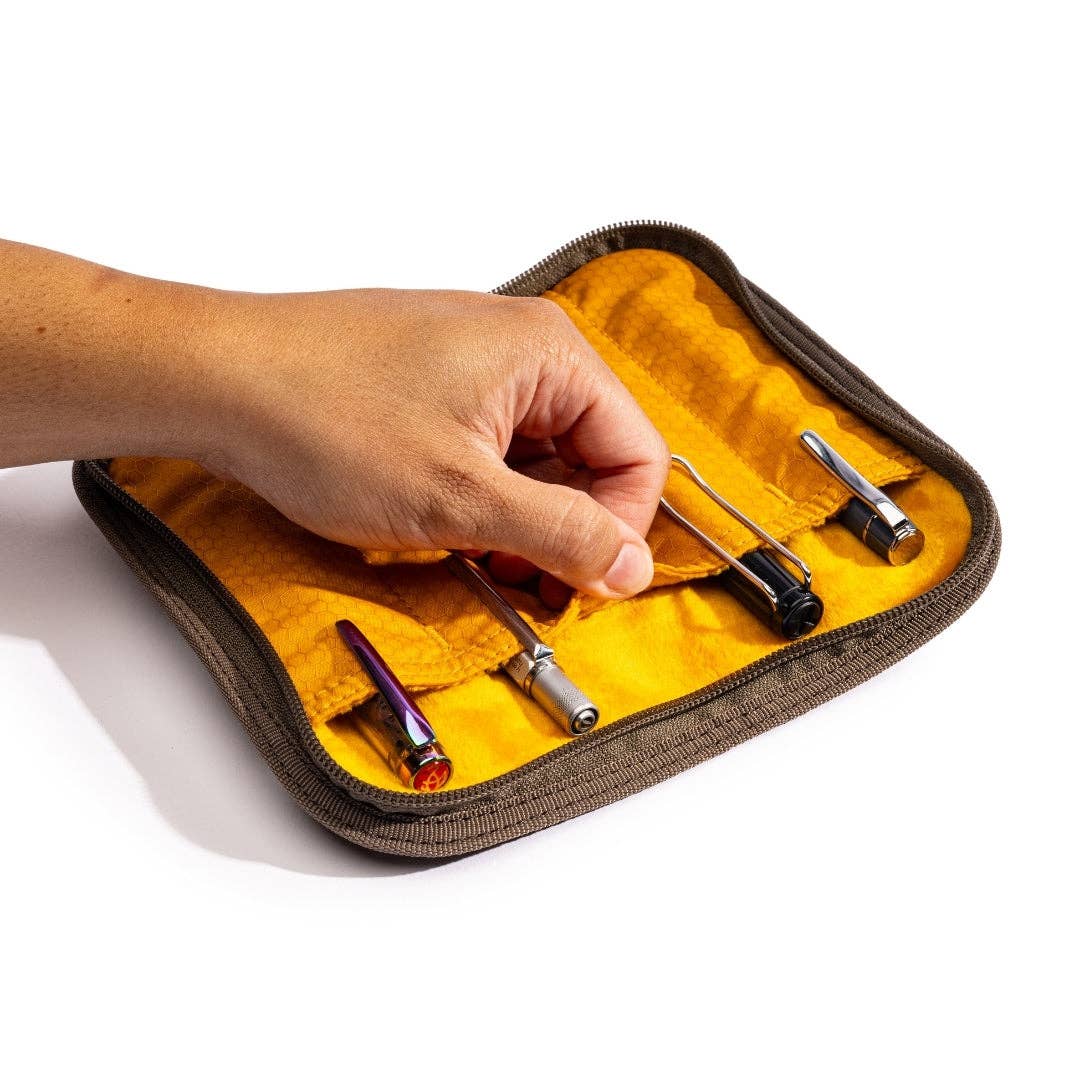 Hand reaching into a small, open pen case with yellow interior containing metallic tools on a white background.