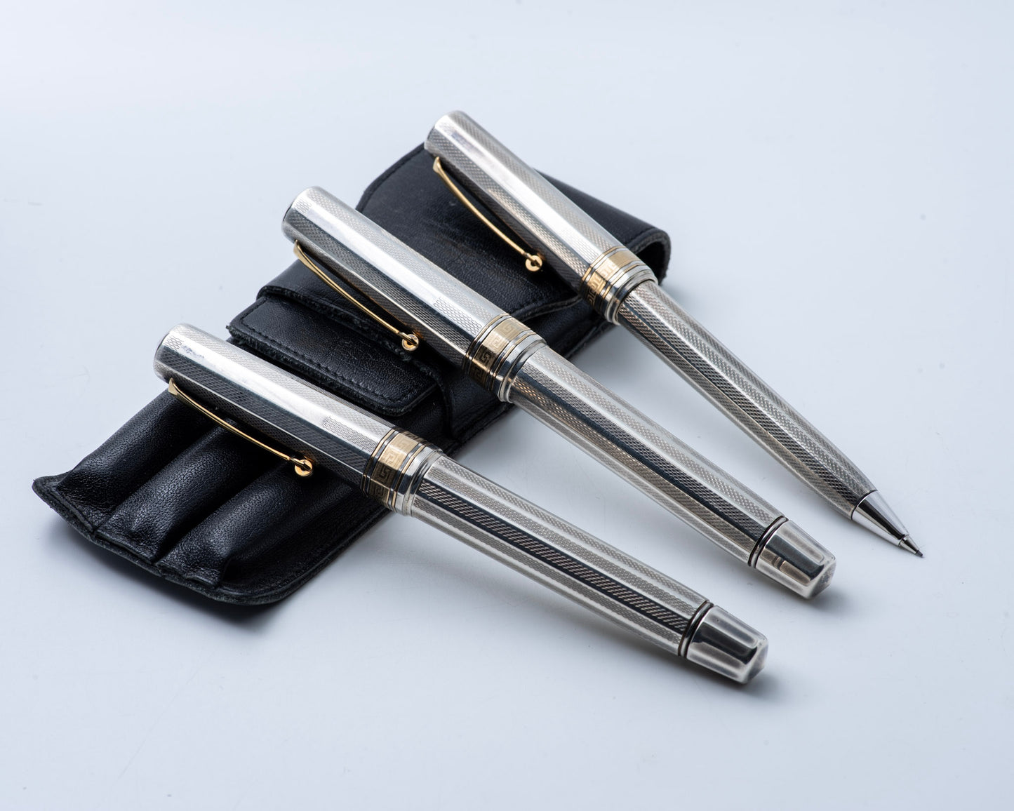 Set of three silver pens with gold accents on a black leather case against a light gray background