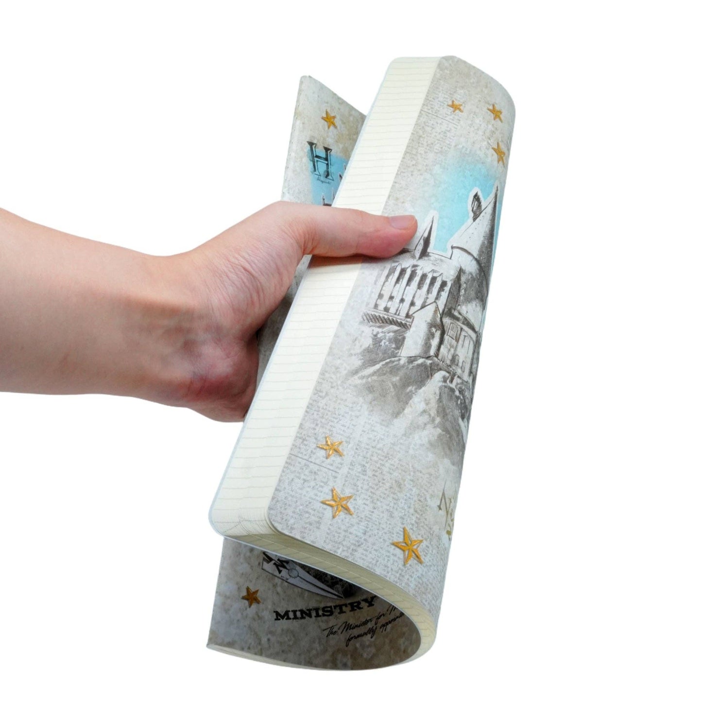 A hand holding a rolled Paperblanks_Harry_Potter_Wizarding_World_Softcover_Cahier notebook featuring a gray and white castle illustration, gold stars, and a light blue sky design on a plain white background.