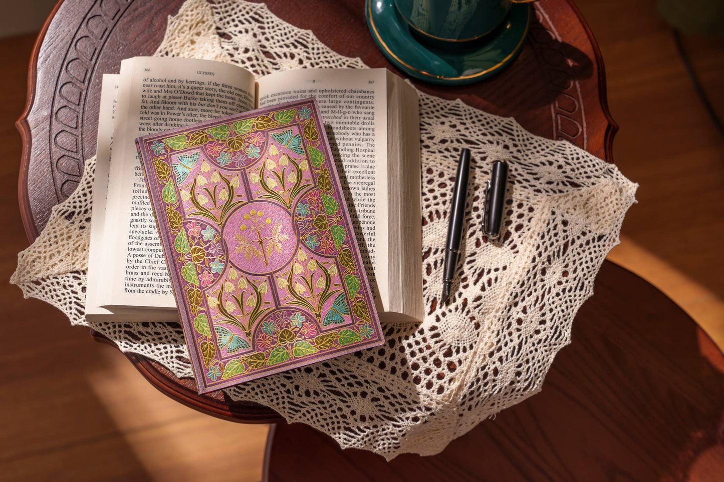 Open book with a colorful cover on a wooden table with a teacup and two pens.