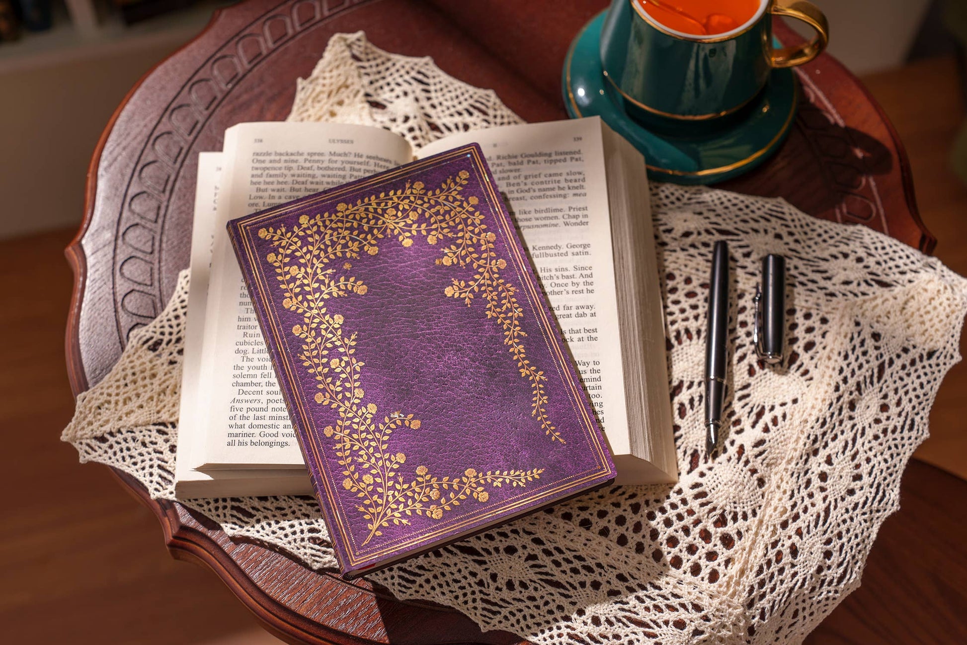 Open book with decorative purple cover on a wooden table, accompanied by a cup and saucer.