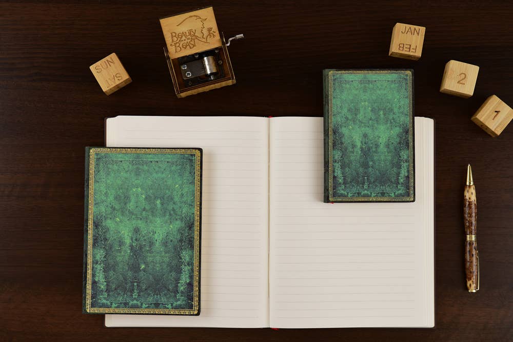 A styled flat-lay of a dark wood desk featuring a vintage-style green textured notebook, a matching green textured box, an open lined notebook, a wooden hand-crank music box, wooden calendar blocks, and a gold pen.