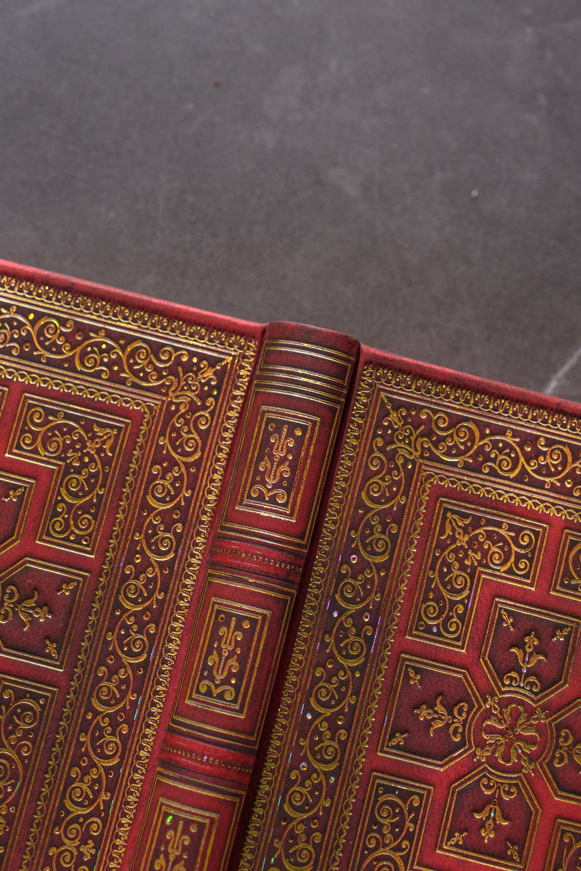 Close-up of an ornate red leather-bound book with intricate gold embossed detailing on the cover and spine, photographed against a dark gray textured background.