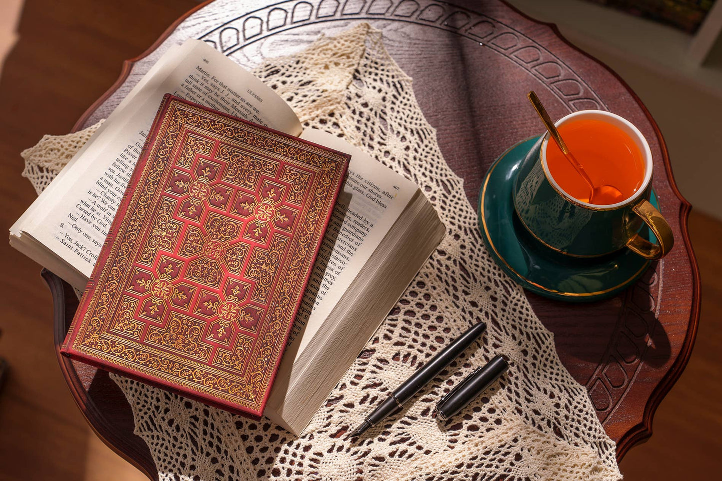 A decorative red and gold hardcover book resting on an open book on a white lace doily, with a teal cup of tea, a spoon, and two black pens on a round wooden table.