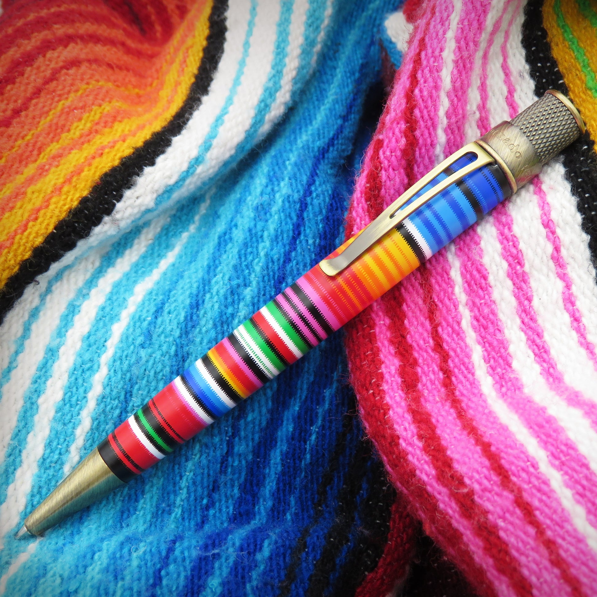 A colorful striped poncho-like pen with a gold-toned clip and tip, resting diagonally on a vibrant, multicolored knit blanket with blue, pink, red, yellow, and white patterns.