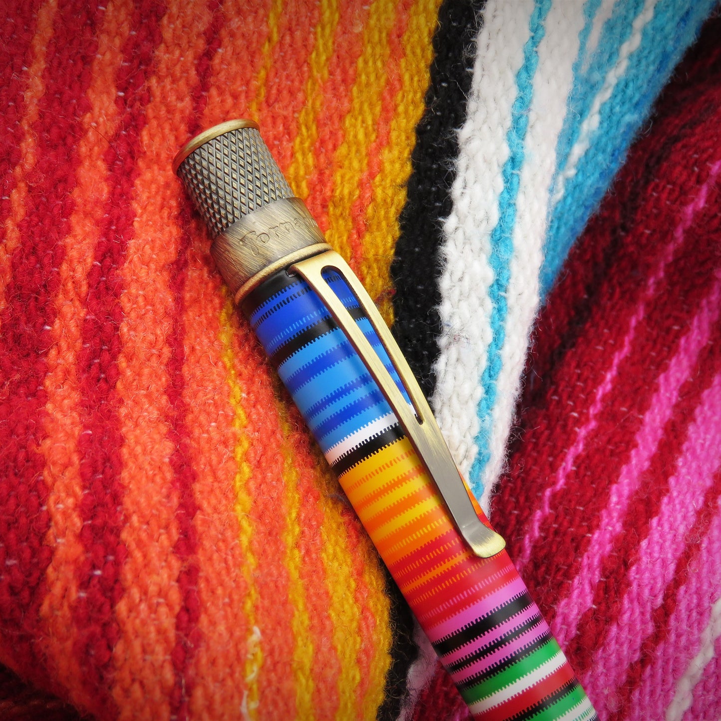 A close-up of a Retro 51 Serape or Poncho pen with a colorful striped handle and a gold-toned clip, resting diagonally on a vibrant, multicolored striped textile background.