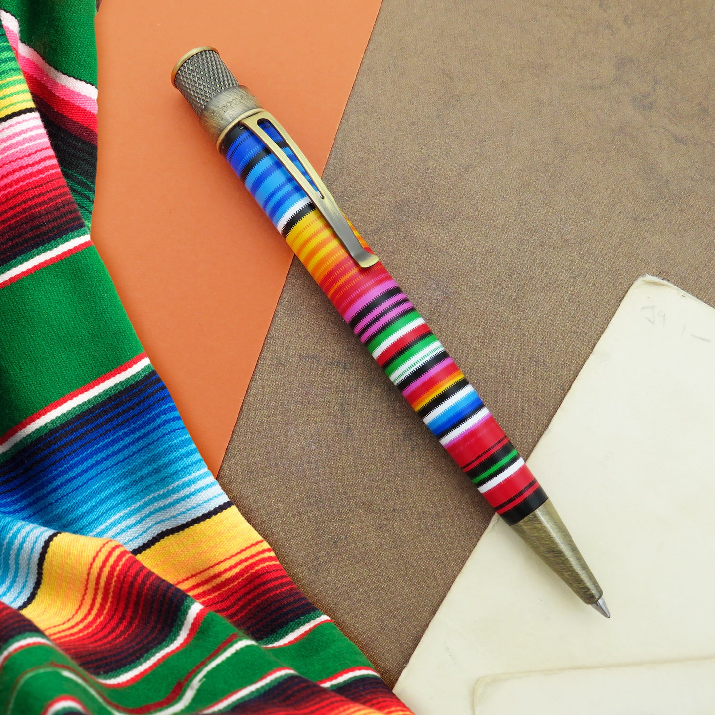 Retro 51 Sarape decorative ballpoint pen with a colorful striped handle, resting diagonally on a brown surface next to a green and red patterned textile.