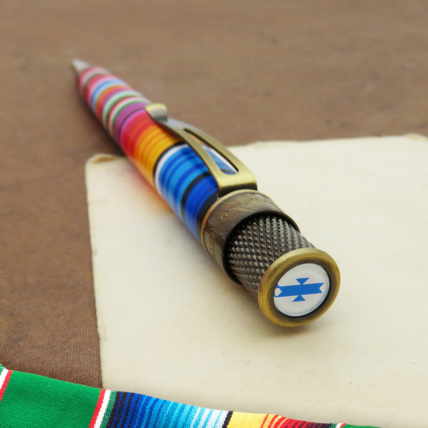 A close-up of a poncho pen resting on a cream-colored card over a brown textured surface. The pen has a colorful striped handle, a gold-toned clip, a dark textured grip, and a round gold-toned end cap with a white circular emblem featuring a blue cross and ribbon.