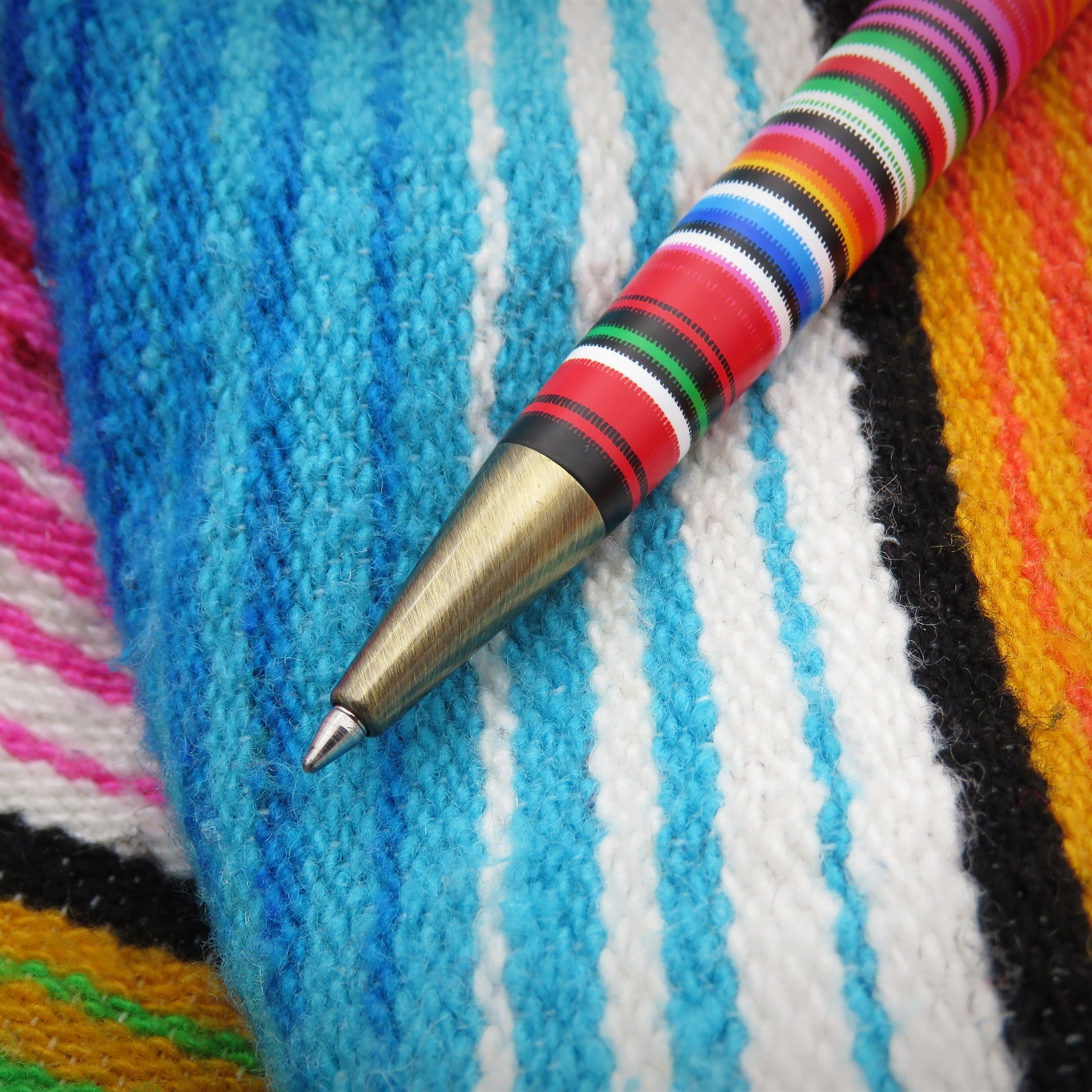 A close-up of a poncho-like pen with a gold-toned metal tip resting on a colorful knit blanket with blue, white, pink, black, and yellow stripes.