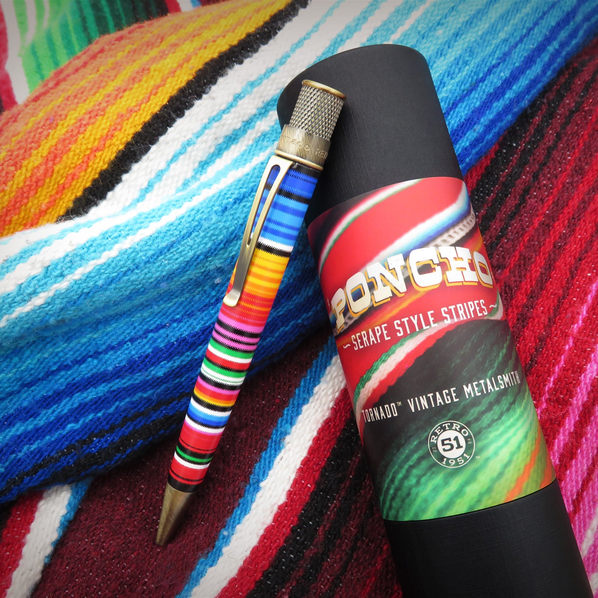 A poncho-striped metal pencil with a gold clip rests on top of its black cylindrical packaging, which features a colorful label. The background is a vibrant, multicolored woven textile with horizontal stripes.