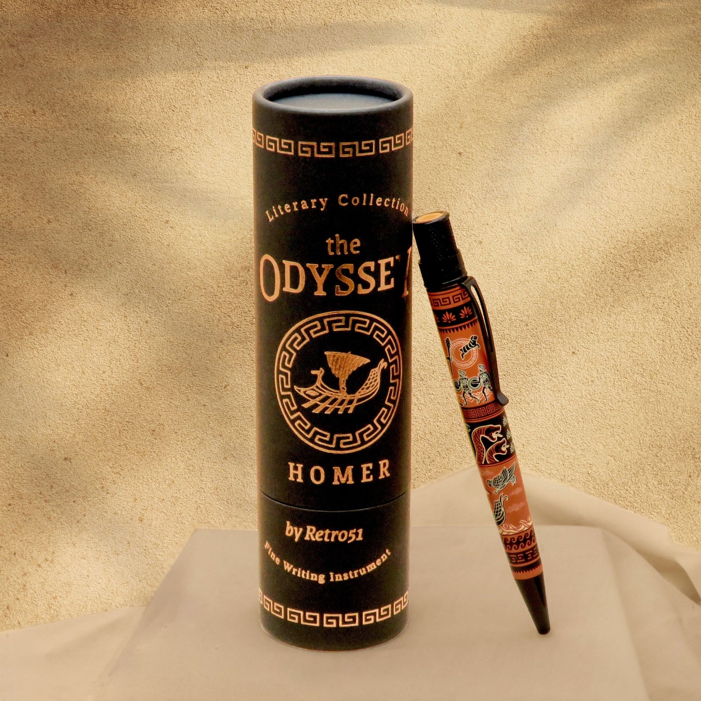 A black cylindrical tube labeled 'the ODYSSEY HOMER' stands upright on draped white fabric, with a matching decorative pen resting beside it against a warm beige textured background.