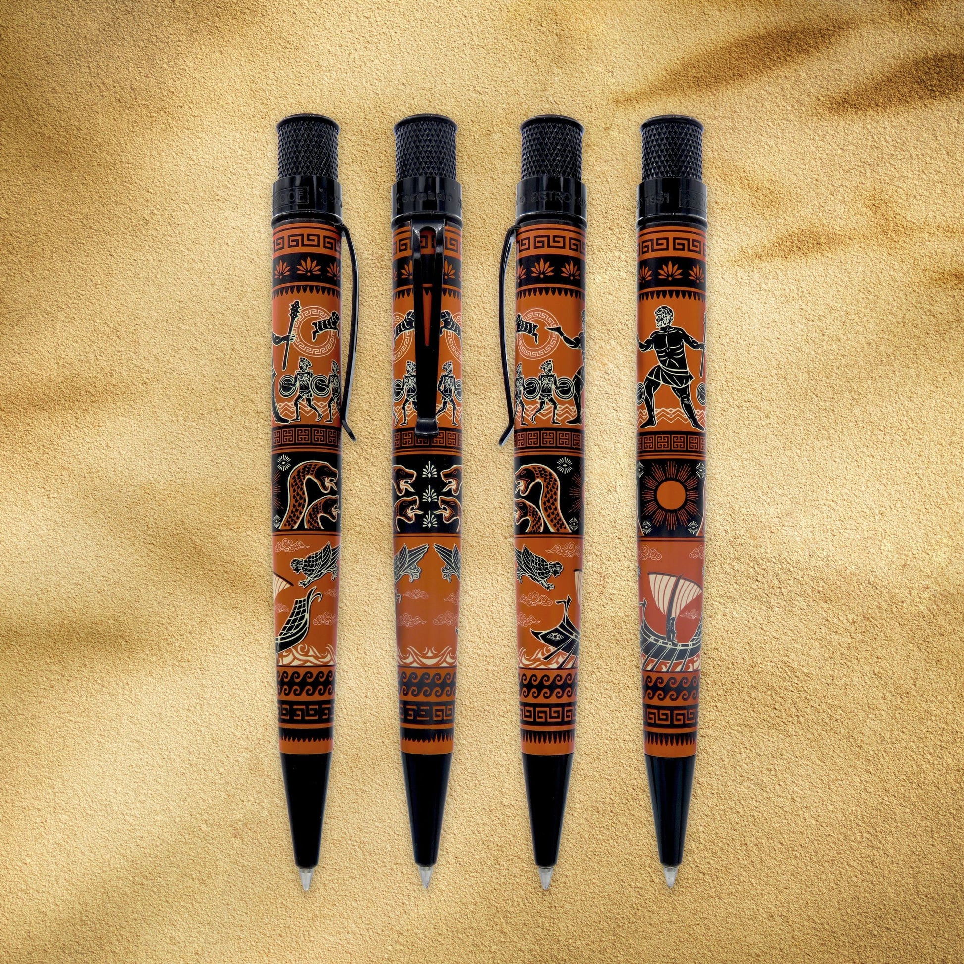 Four decorative ballpoint pens with black caps and tips, featuring an orange and black tribal-style pattern, arranged vertically on a textured beige background.