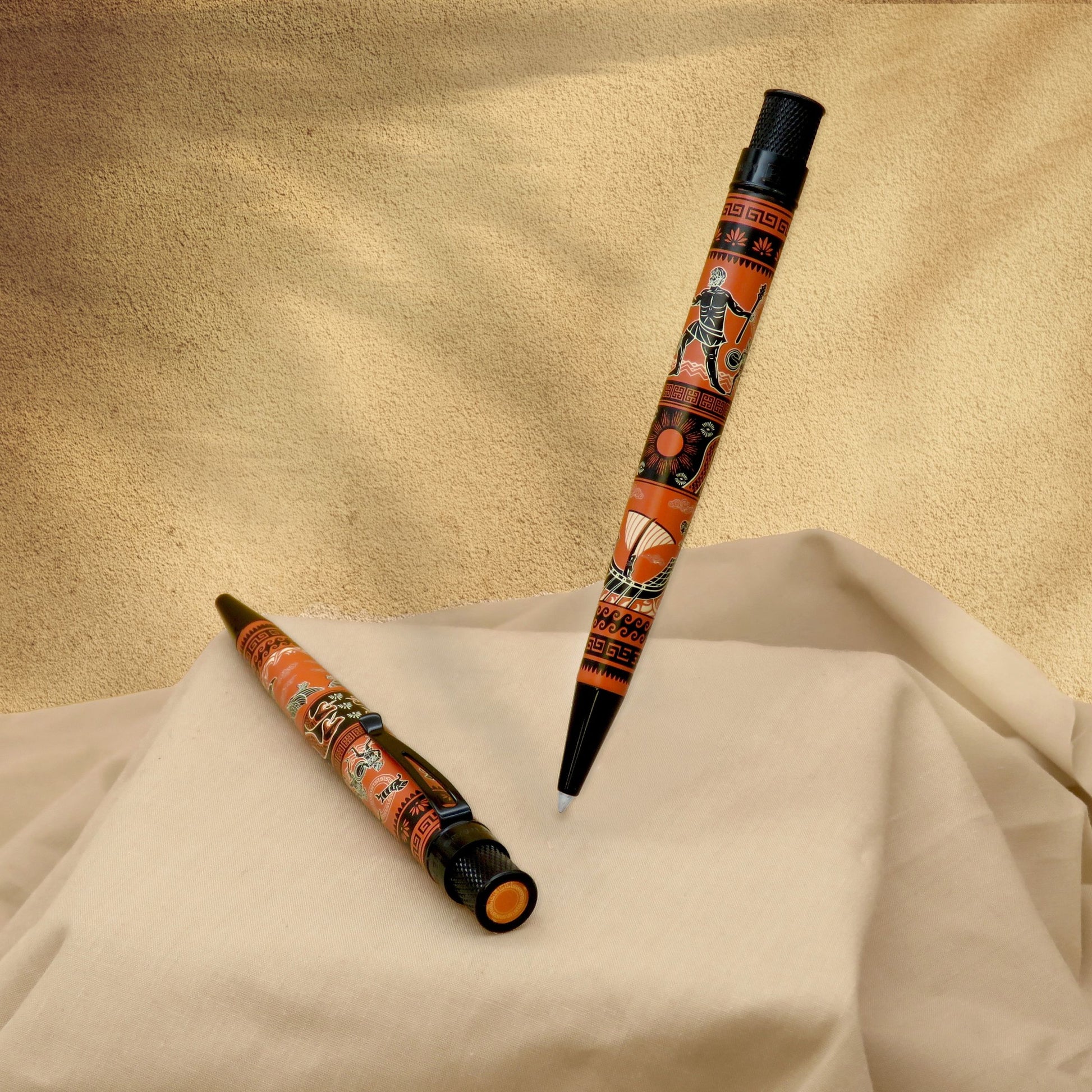 Two decorative ballpoint pens with an orange, black, and brown Aboriginal-style pattern, displayed on draped cream fabric against a textured beige background.