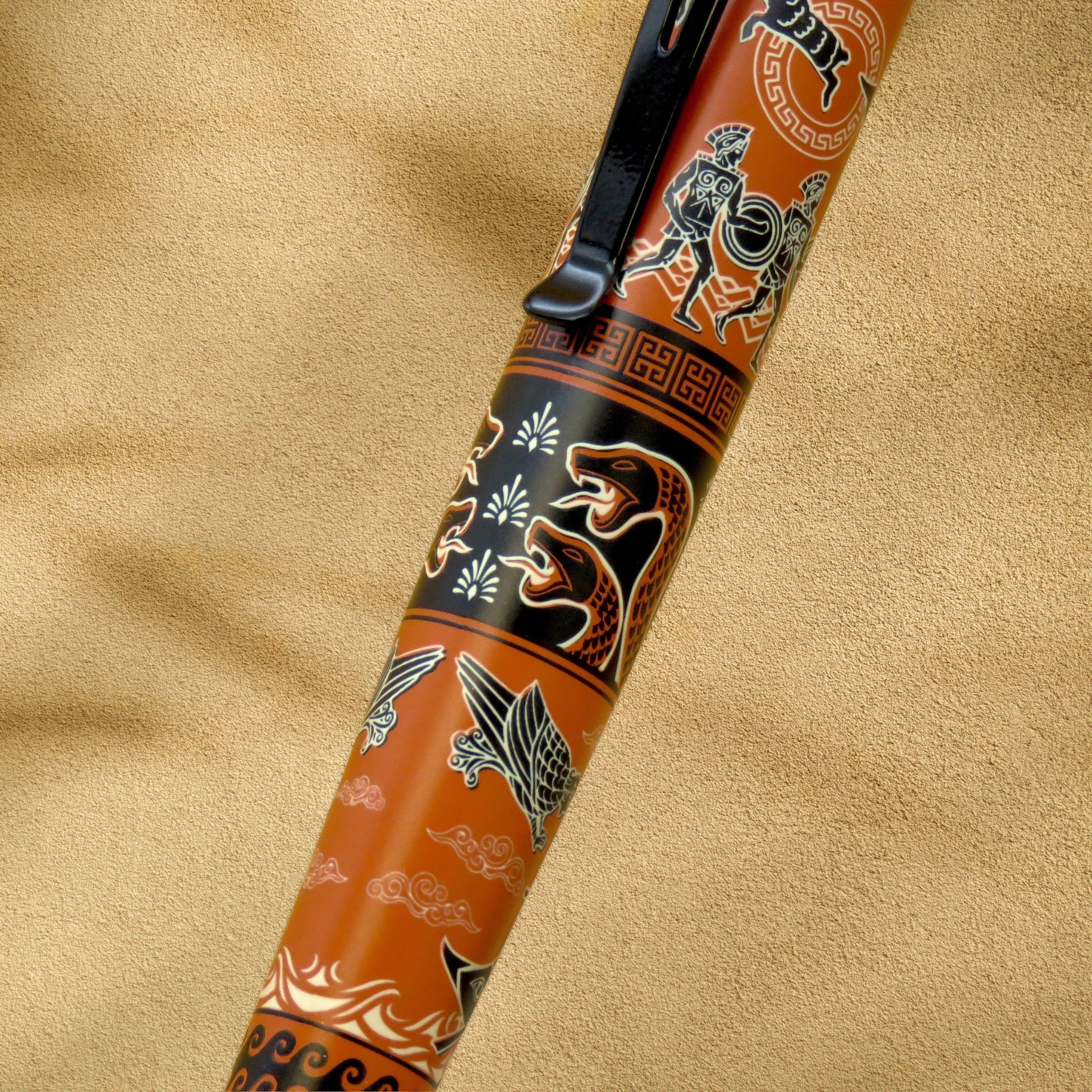 A close-up of a decorative pen with an orange body, black clip, and detailed black, white, and gold traditional-style illustrations, set against a textured beige background.