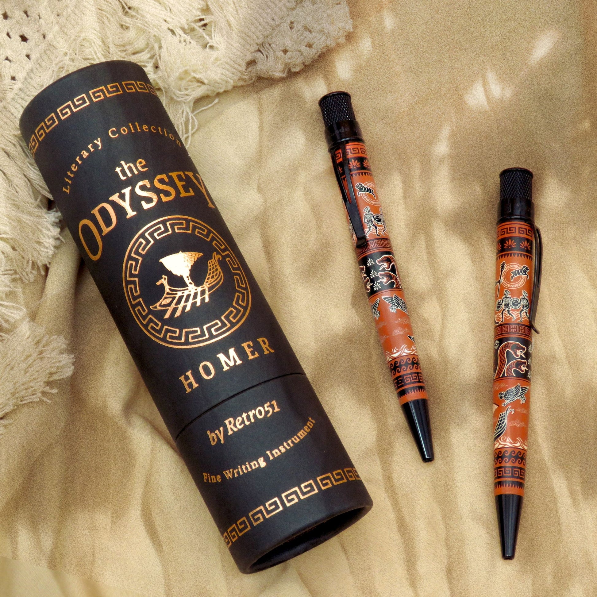 A black cylindrical presentation tube labeled 'The Odyssey Homer' by Retro51, shown beside two matching orange-and-black pen-style writing instruments on a beige fabric background.