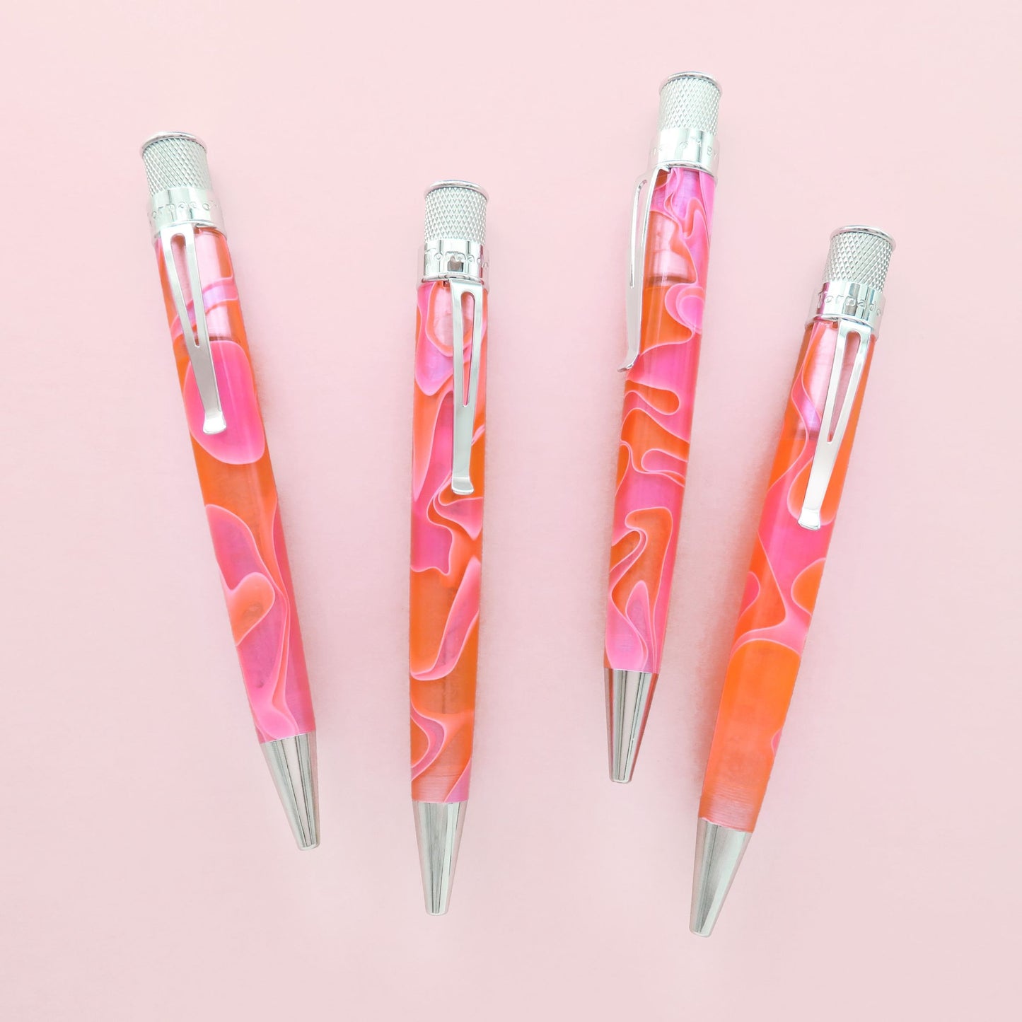 Four  lava-like pens with a pink and orange marbled pattern, silver clips, and silver tips, arranged diagonally on a solid pink background.