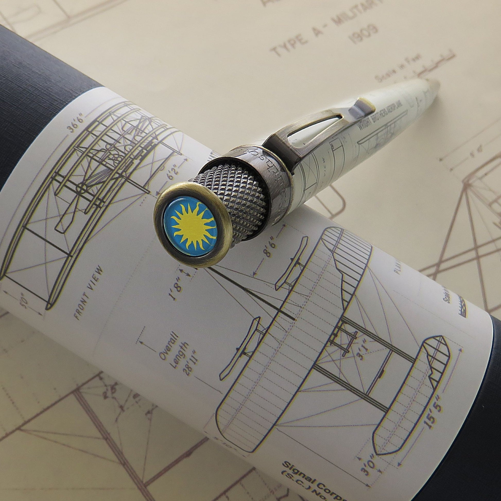 A close-up of a Retro 51 Wright Brothers Aeroplane - Rollerball pen resting on a rolled blueprint, featuring a circular gold-toned end cap with a blue background and yellow sun emblem.