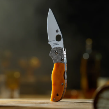 A Spyderco Native 5 50th Anniversary folding pocket knife with a silver blade and an orange handle, standing upright on a wooden surface with a blurred dark background.